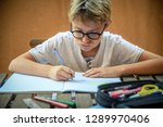 Small photo of Cute blond child with glasses sitting at the table doing homework for school Young student intent on studying Boy concentrated in the study Fatigued and bored, snort Writing on an exercise book