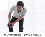 Small photo of Asian old middle aged senior male grandfather wears white shirt black pants stand bend down hold his hands on right leg show moody face when has pain and knee ache alone in front clear white curtain.