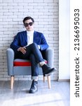 Small photo of Studio shot Asian young smart handsome successful professional male businessman in casual blue suit turtle neck shirt sunglasses sit on armchair looking at camera on brick wall background