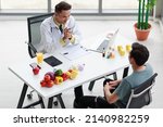 Small photo of Top view shot Asian male professional nutritionist dietitian doctor in white lab coat and stethoscope sitting smiling discussing with patient at working desk about nutrition food and supplement pill.