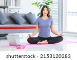 Small photo of Asian young happy peaceful calm female model in casual sporty outfit sitting crossed legs in lotus position on yoga mat learning studying online meditation class via laptop computer in living room.