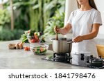 Photo of pretty caucasian woman holding spoon while cooking soup with fresh vegetables in open kitchen at home in Bali