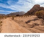 Small photo of A sandy trail winds through a sparse desert landscape near Tafraout Morocco, with a small rocky hill rising amidst scant vegetation, backed by distant mountains