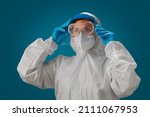 Small photo of A female doctor or nurse wearing a PPE suit, white N95 mask, goggles, and blue medical gloves puts on a visor as a precaution. Personal protective equipment to protect against coronavirus covid-19.