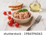 Canned tuna meat in a bowl, fork, bread and fresh red cherry tomatoes on a white wooden table. Low calories healthy eating snack of preserved tuna fish and vegetables. Tasty seafood. Front view.
