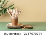 Disposable dishes and utensils against beige green background. Biodegradable wooden cutlery in brown paper cups on plates. Eco-friendly tableware and recycling concept. Copy space. Front view.
