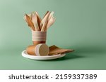 Stack of eco-friendly disposable tableware. Wooden forks and knives, paper cups and plates against green background. Biodegradable cutlery and dishes for picnics, takeaways. Copy space. Front view.
