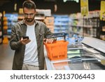 Small photo of At the Supermarket: Handsome Man Uses Smartphone and Looks at Nutritional Value of the Canned Goods. He's Standing with Shopping Cart in Canned Goods Section