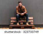 Confident and handsome. Handsome young man sitting on the wooden pallet and looking at camera with grey wall in the background and orange fallen leaves on the floor 