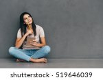Waiting for inspiration. Attractive young woman in casual wear holding notebook and pen while sitting barefoot and against grey background