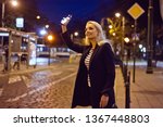 Young blonde woman standing in the city street in the evening, holding smart phone in hand and waiting for taxi or uber.