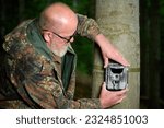Small photo of Hunter sets a trail camera on a tree in the forest. Trail cameras are often used by hunters for automatic photography or video shooting of wildlife in the forest.