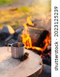 Small photo of Metal campfire enamel mug with hot herbal tea on wooden stump. Bowler pot and bonfire on background, cones. Concept of lunch break during hiking, trekking, active tourism, camping