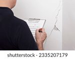 Small photo of Rental damage concept: man with german inspection checklist - translation: protocol of tenancy changover - in front of a white wall with a long crack or rip.