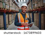 Happy male factory manager using digital tablet in warehouse while standing against goods shelf looking at camera