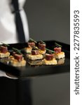 Small photo of The waiter holds a Set of assorted festive appetizer canapes on bread on a black tray as an event dish. Buffet table. Catering. Delicacy. Haute cuisine.