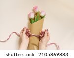 Flat female hands tie a satin ribbon bow on a simple bouquet of fresh pink tulip flowers in craft paper on a beige table.