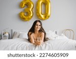 Small photo of Happy young woman meets her thirtieth birthday in bed, holding a glass of champagne in her hands and smiling