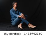 Black young beautiful happy smiling lady wearing jeans sitting on the floor with crossed legs and shoes against black background
