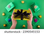 Dive into holiday magic! First person top view of hands reach for lavish golden giftbox featuring ribbon bow near green gift boxes surprise on green backdrop. Perfect for Black Friday marketing
