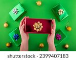 Dive into holiday magic! First person top view of hands reach for lavish red giftbox featuring ribbon bow near green gift boxes surprise on green backdrop. Perfect for Black Friday marketing