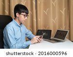 Small photo of Happy young Asian businessman or investor using smartphone for trading, investing on new cryptocurrency or stock trading platform at laptop, tablet with stock tickers and graphs. Young money concept.