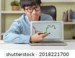 Small photo of Happy young Asian businessman or investor pointing on trading tickers or graphs on laptop to teach online investing courses on stock market or Cryptocurrency as Bitcoin at home. Young money concept.