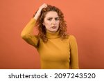 A young and attractive Caucasian redhead girl in an orange jumper holding her hand on her head because she is confused isolated on an orange studio background.