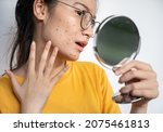 Small photo of Young Asian woman feeling shock while looking acne and scar occur on face by mini mirror. Conceptual shot of Acne and Problem Skin on female face.