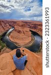 Small photo of Tourist man sitting at the cliff edge with panoramic aerial view of Horseshoe bend on the Colorado river near Page in summer, Arizona, USA. Incised meanders in Glen Canyon National Recreation Area