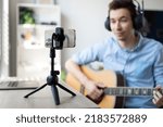 Blurred shot of young man in headset sing song play guitar streaming at real time on personal music channel at social media. Focus on modern mobile phone fixed on tripod holder.