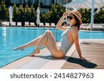 Woman in striped swimsuit, hat and sunglasses sitting by the pool. The concept of summer luxury vacation in the hotel