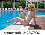 Woman in striped swimsuit, hat and sunglasses sitting by the pool. The concept of summer luxury vacation in the hotel