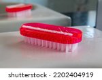 Small photo of A red nail brush with white bristles on a white ceramic sink in a domestic bathroom with a mirror behind, narrow depth of field focus on the nearer bristles