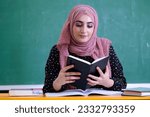 Teacher or student wearing Hijab scarf inside classroom with blackboard as background for education, university campus concept. Female holding a notebook at school