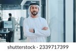 Small photo of Emirati Arab at office wearing Kandura looking at front ideal for Middle East business concept. Arabic man inside a corporate establishment with colleagues at the background.