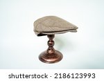 Small photo of Detail of scally cap or flat cap in dark brown pattern herringbone tweed fabric set on a bronze head mannequin on a white background