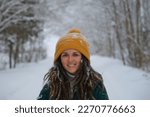 Happy European girl with blue eyes and dreadlocks walks alone in winter park. Close up portrait of young smiling pretty Caucasian woman in yellow knitted hat against background of snowy winter forest.