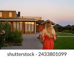 A girl with blond hair with her back to the camera walks into a country house at sunset