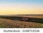 Tractor Plowing field on sunset. Red Tractor with Plough on Plowed. Ploughing and Soil Tillage. Agricultural Tractor on Cultivation Field for Sowing Seeds. Big Tractor During Field Cultivating. 
