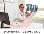 Small photo of Serious pulmonologist comparing two x-ray pictures of lungs. Young African American doctor sitting at table examining x-ray radiography in her office. Radiology research, pneumonia or asthma concept