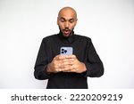 Small photo of Portrait of astounded young man reading message on mobile phone against white background.Bearded businessman wearing black shirt surfing Internet on smartphone. Good news and mobile technology concept