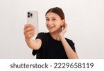 Small photo of Portrait of happy young woman smarten up while looking at phone over white background. Caucasian woman wearing black T-shirt taking selfie or using mirror app. Mobile technology concept