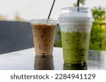Selective focus of iced matcha with white foam milk on top and iced espresso, Takeaway homemade cold drink in plastic glass and straw on the table with blurred green garden background.