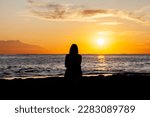 early morning coastal view at sunrise across ocean towards horizon with clouds in colorful sky in background and silhouette of meditating person in dark foreground and sun to the right