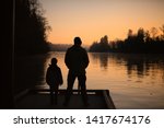 A man and child dark silhouette on a dock watching the orange sunrise on Willammette River, in the cold orange morning light of Wilsonville Memorial Park, Oregon, USA