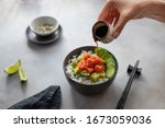 woman's hand pours soy sauce in a bowl with salmon, rice, avocado and cucumber. Food concept poke bowl. horizontal image, gray concrete background.