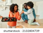 Small photo of An African American family with their little daughter with curly fluffy hair made cupcakes together in the kitchen. Mom brought a baking sheet with ready-made muffins and gives the family a try.