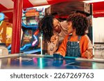 Small photo of A cute African-American child with afro curls with her mother playing air hockey at an amusement park and carousel on her day off in the evening.