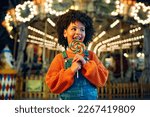 A cute African-American girl with an Afro hairstyle eats a colorful lollipop standing against the background of a carousel with horses in the evening at an amusement park or circus.